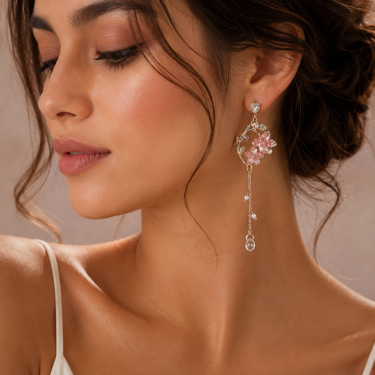 A close-up of a woman wearing a dangling earring with pink flower-shaped crystals and silver-toned chain drops against a soft beige background.