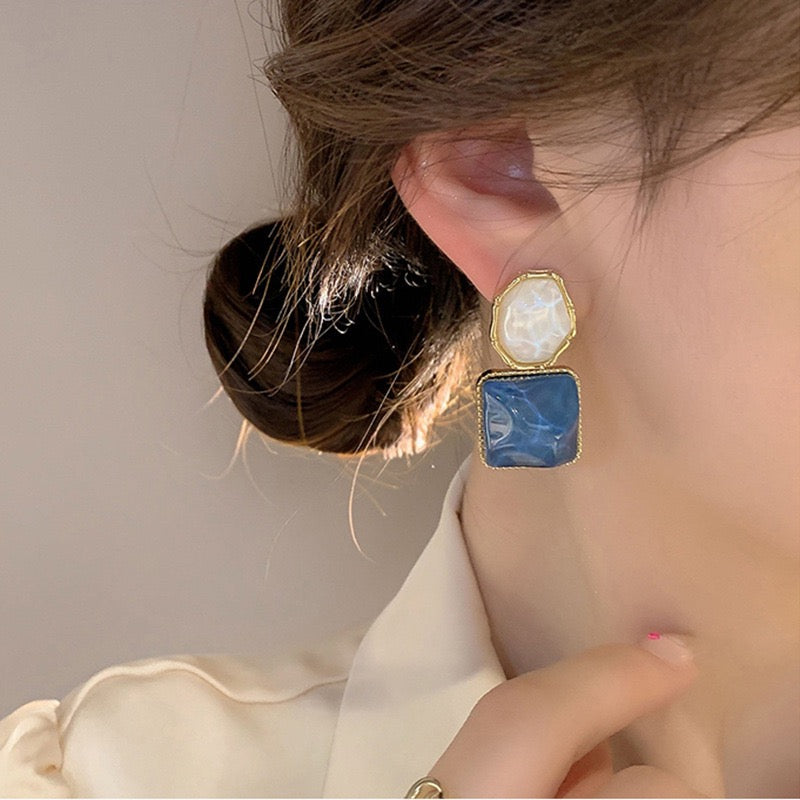 A close-up of a person wearing a gold-tone double-stone earring with a textured cream oval top and a square blue bottom stone.