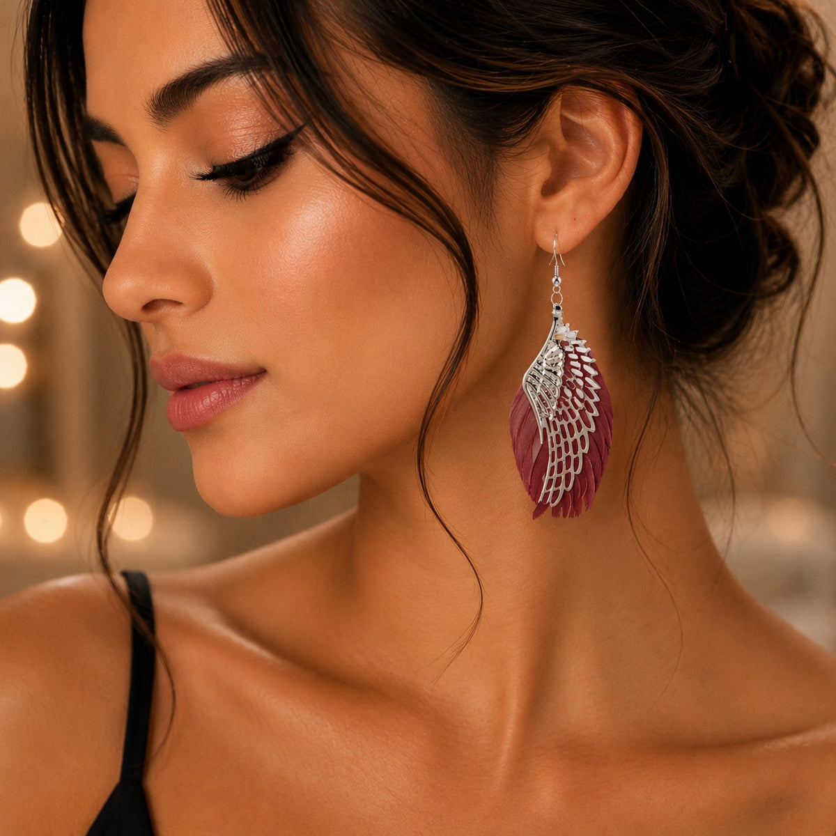 A close-up lifestyle shot of a woman wearing a dangling feather earring. The earring features a silver-toned metal filigree top and a burgundy-pink feather drape below, shown against a warm, softly blurred background with bokeh lights.