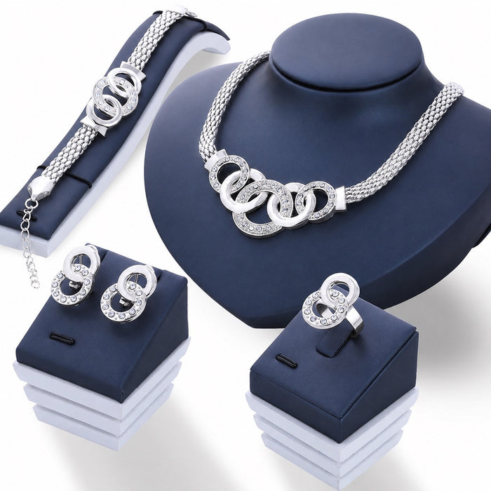 A silver-tone jewelry set displayed on dark blue stands, including a necklace with interlocking ring motifs, a bracelet, a ring, and matching earrings, all embellished with clear rhinestones.