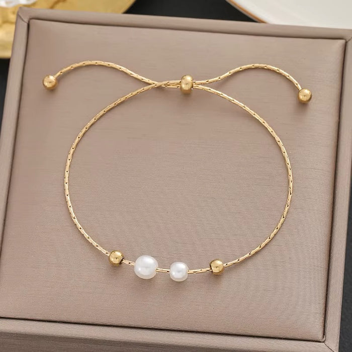 A delicate gold-toned adjustable bracelet with three white pearl-like beads, displayed on a textured taupe jewelry box.