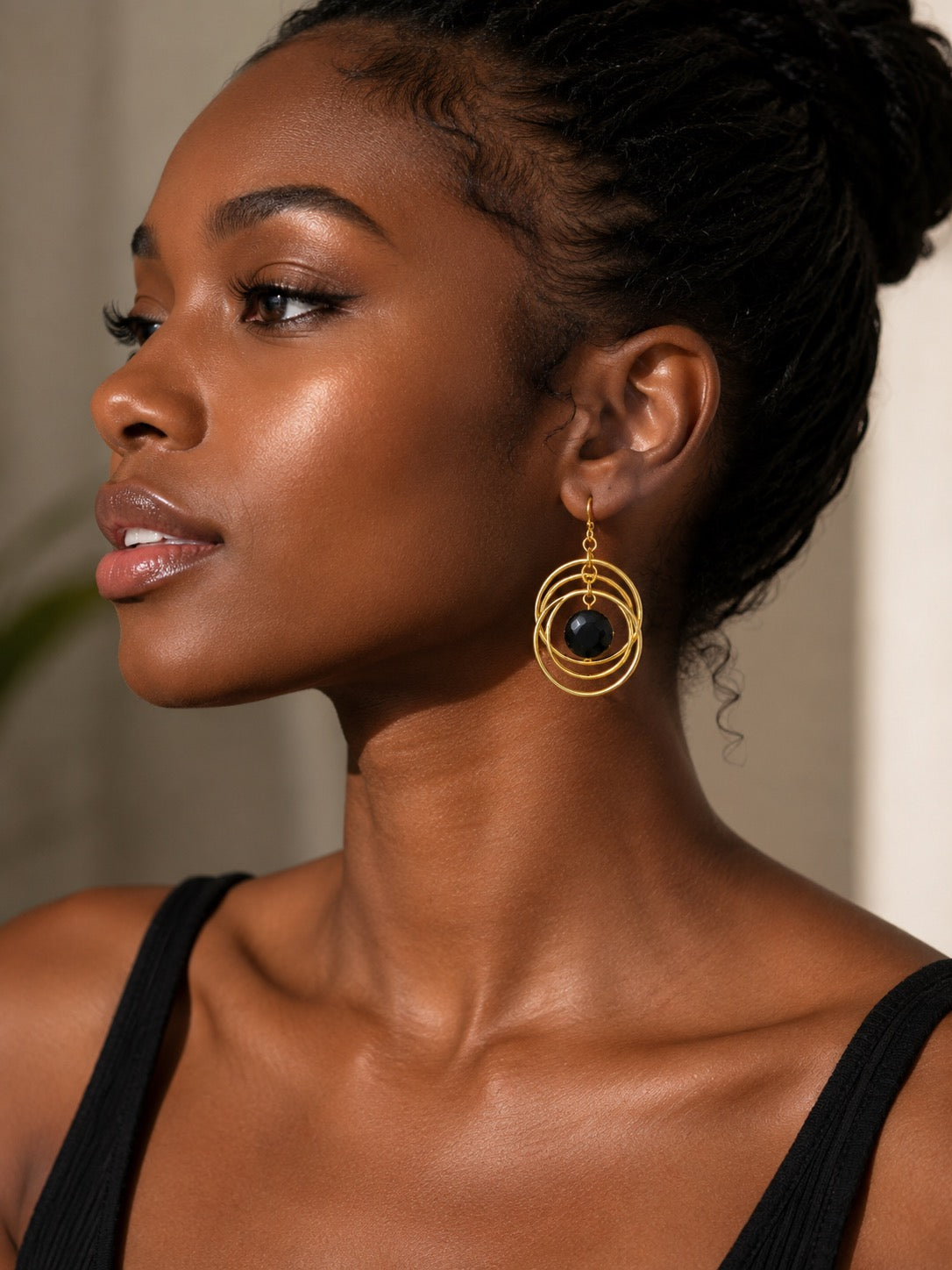 A close-up side profile of a woman wearing a pair of gold-toned dangle earrings with a central dark oval stone, set against a softly blurred neutral background.