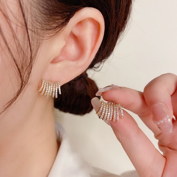Close-up of a person wearing a gold-toned crescent-shaped earring with multiple rows of small clear stones, while holding the matching earring between their fingers against a soft light background.
