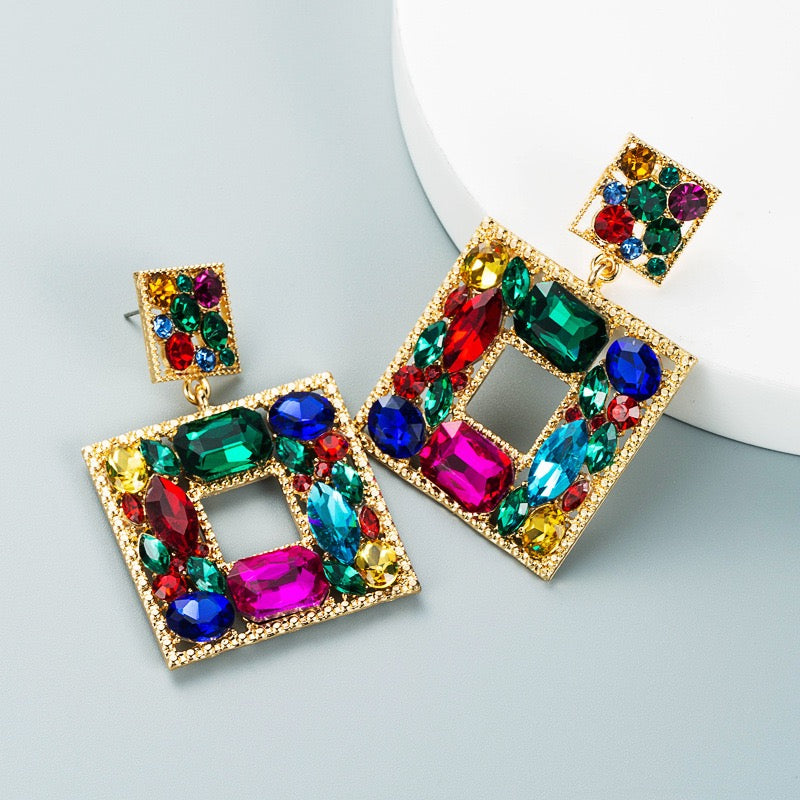 A pair of gold-tone geometric drop earrings adorned with colorful faceted rhinestones in green, blue, pink, red, yellow, and purple, displayed on a light gray surface next to a white curved prop.