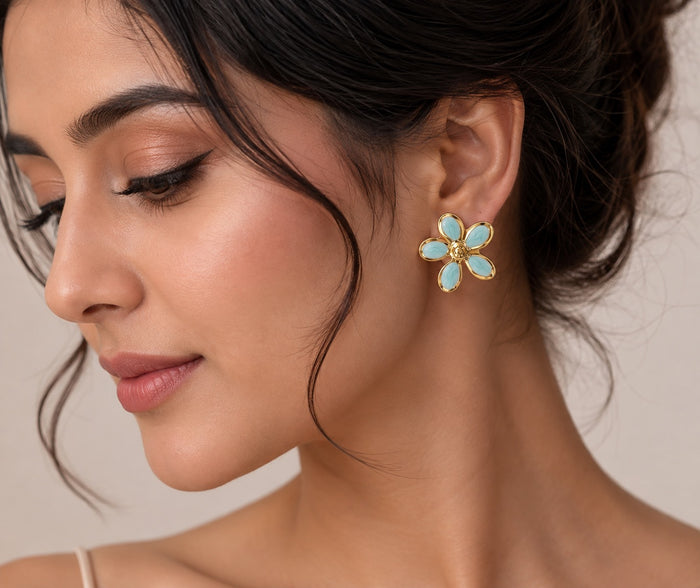 A close-up of a woman wearing a gold flower-shaped stud earring with light blue oval stones, shown against a soft beige background.