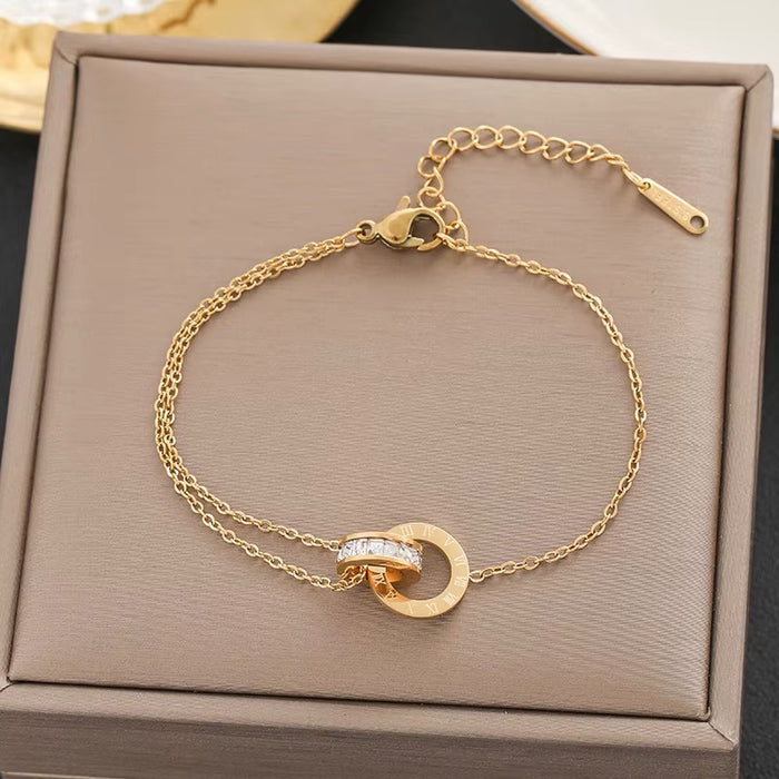 A gold chain bracelet with a double-ring design, one of which is set with small clear stones, displayed on a textured taupe jewelry box.