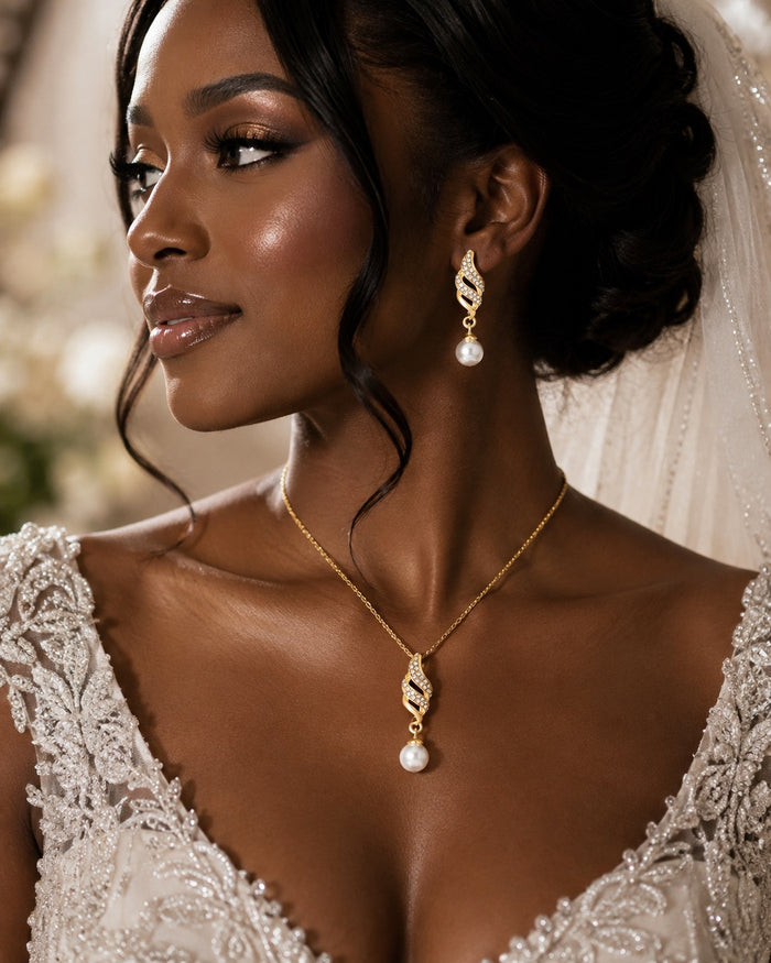 A close-up portrait of a woman in a white lace bridal gown wearing a gold pendant necklace and matching drop earrings with pearl-like beads, against a softly blurred warm background.