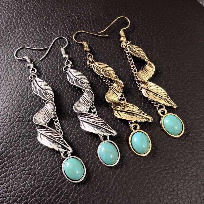 Three pairs of dangle earrings displayed on a dark brown textured leather background. The earrings feature oxidized silver and gold leaf designs with oval turquoise-colored stone pendants.