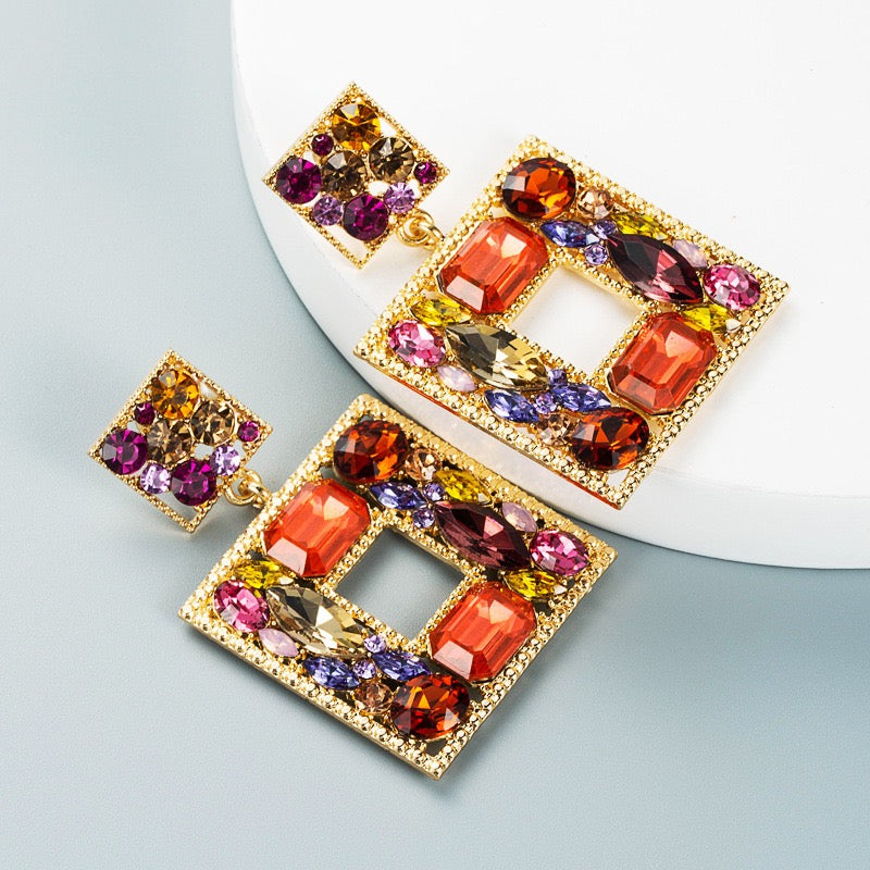 A pair of gold-tone statement earrings with square dangling charms, embellished with multiple colorful gemstones including orange, pink, purple, yellow, and blue, displayed on a white curved platform over a light blue-gray background.
