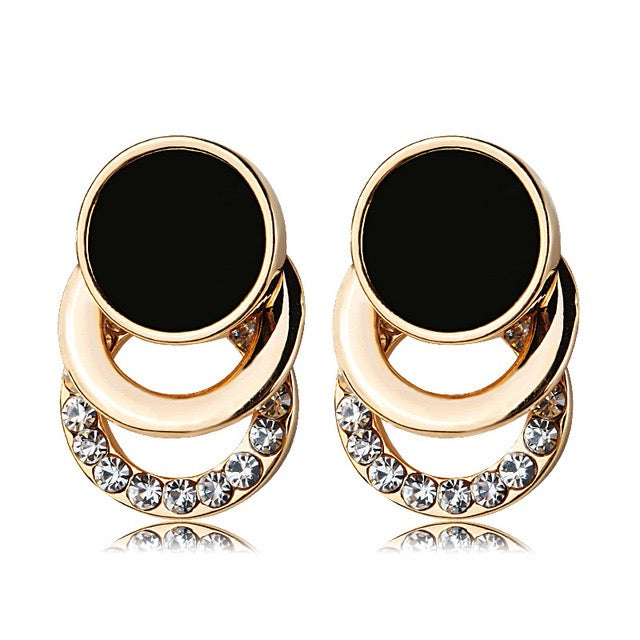A pair of gold-plated stud earrings featuring a large black circular disc, a smaller gold disc, and a curved lower section set with clear rhinestones, shown on a white background with a soft reflection.