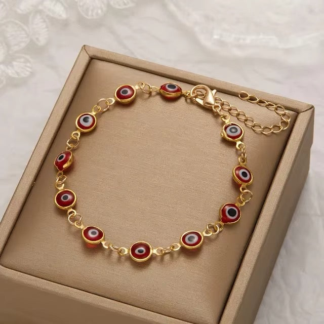 A evil eye charm bracelet with red enamel and white, black, and blue details, displayed inside a beige jewelry box on a white lace fabric background.