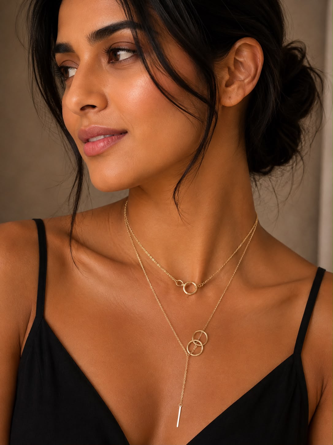 A close-up of a woman wearing a delicate gold layered necklace with two interlocking rings and a long Y-shaped drop ending in a thin bar, paired with a black spaghetti-strap top.