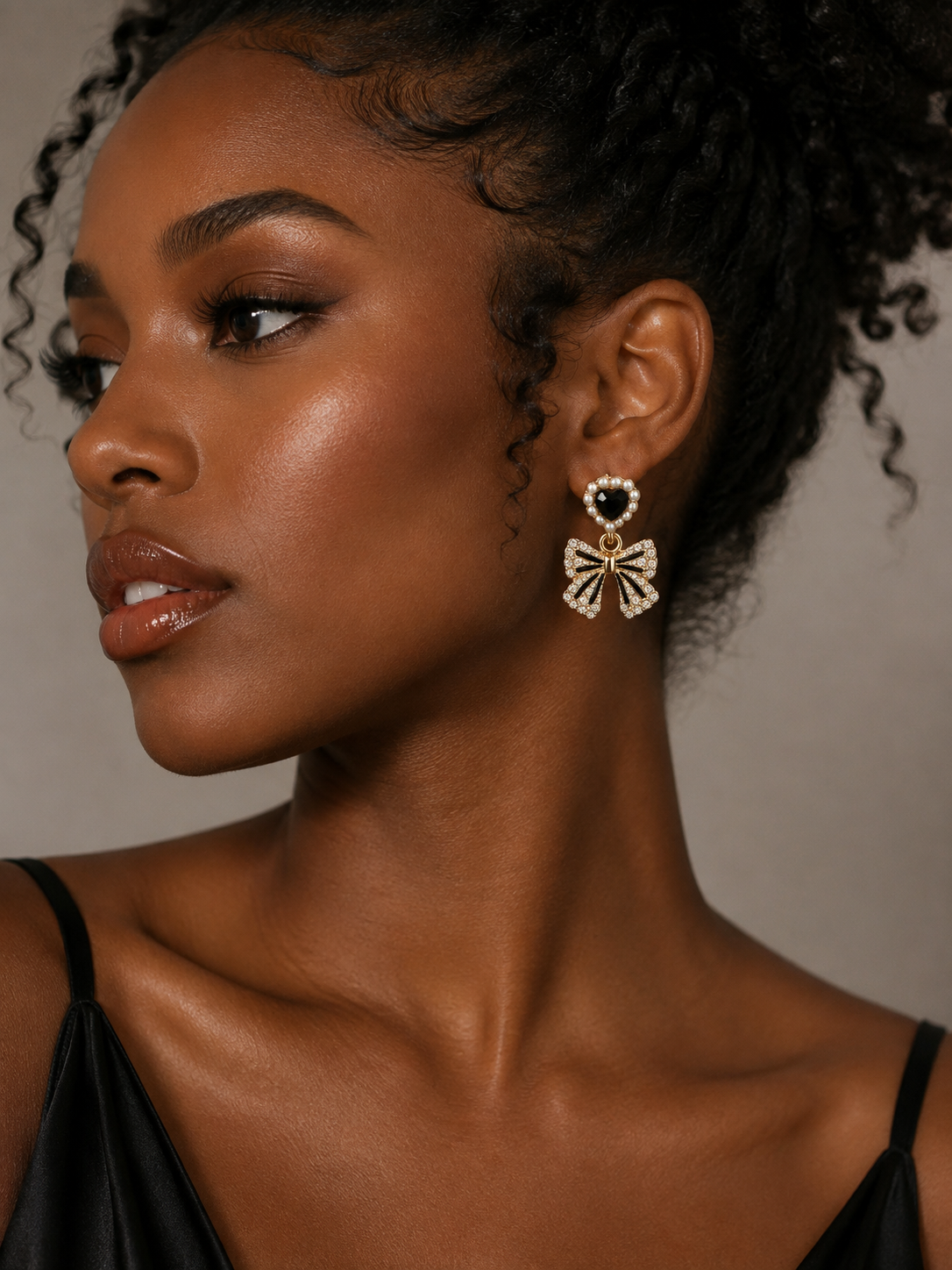 A close-up portrait of a woman wearing a dangling bow-shaped earring with a heart-shaped top and small clear stones, shown against a plain warm gray background.
