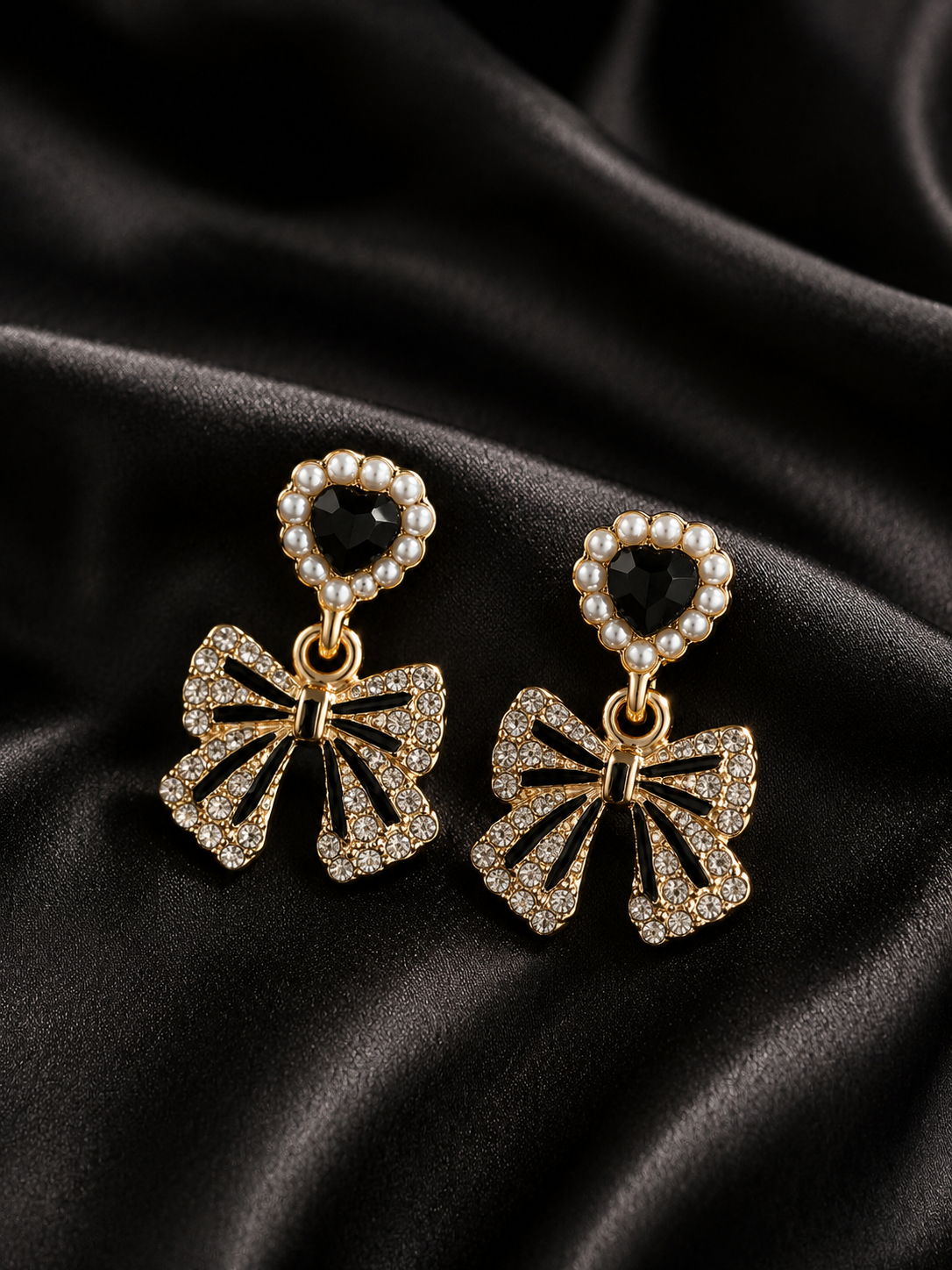 A pair of gold-toned dangle earrings with heart-shaped pearl borders, black heart gemstones, and dangling bow charms encrusted with clear stones, displayed on a draped black satin fabric.