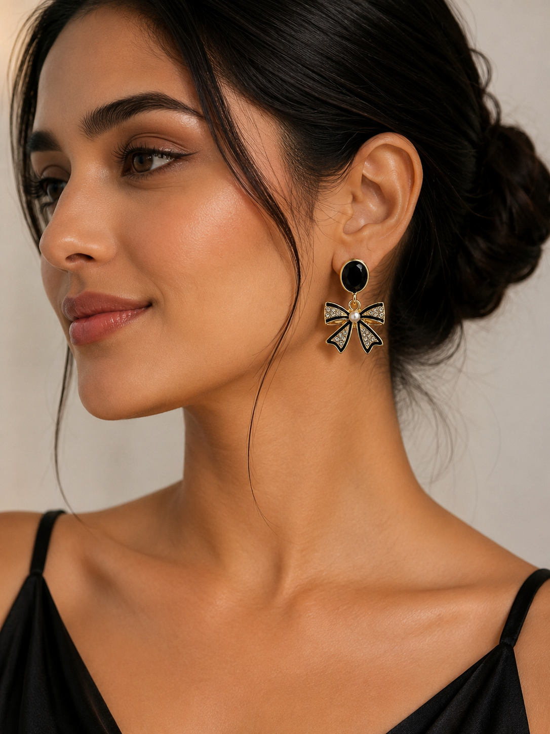 A close-up of a woman wearing a dangling earring with a black oval stone and a bow-shaped charm outlined in gold with small light blue stones, against a soft beige background.
