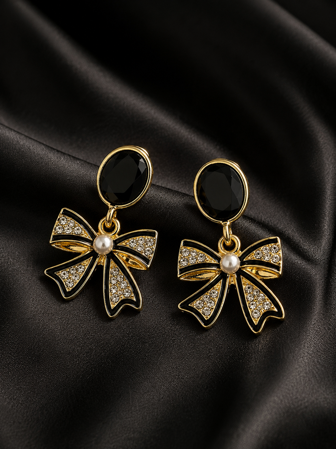 A pair of gold bow-shaped drop earrings with black oval studs, clear rhinestone accents, and a small white pearl at the center, displayed on a draped black satin fabric.