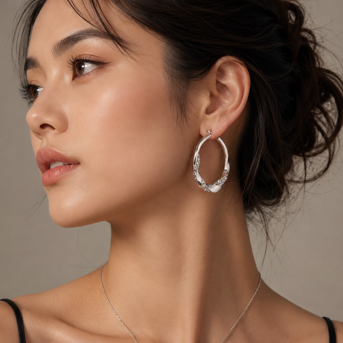 A close-up side profile of a woman wearing a polished silver hoop earring adorned with small clear stones, paired with a delicate silver necklace against a warm neutral background.