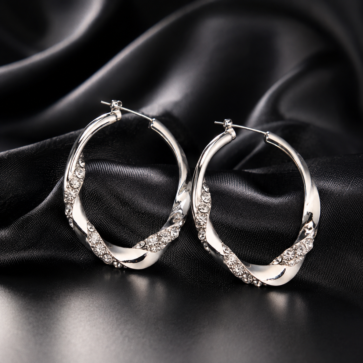 A pair of silver-toned twisted hoop earrings with clear crystal accents, displayed on a draped black satin fabric background.