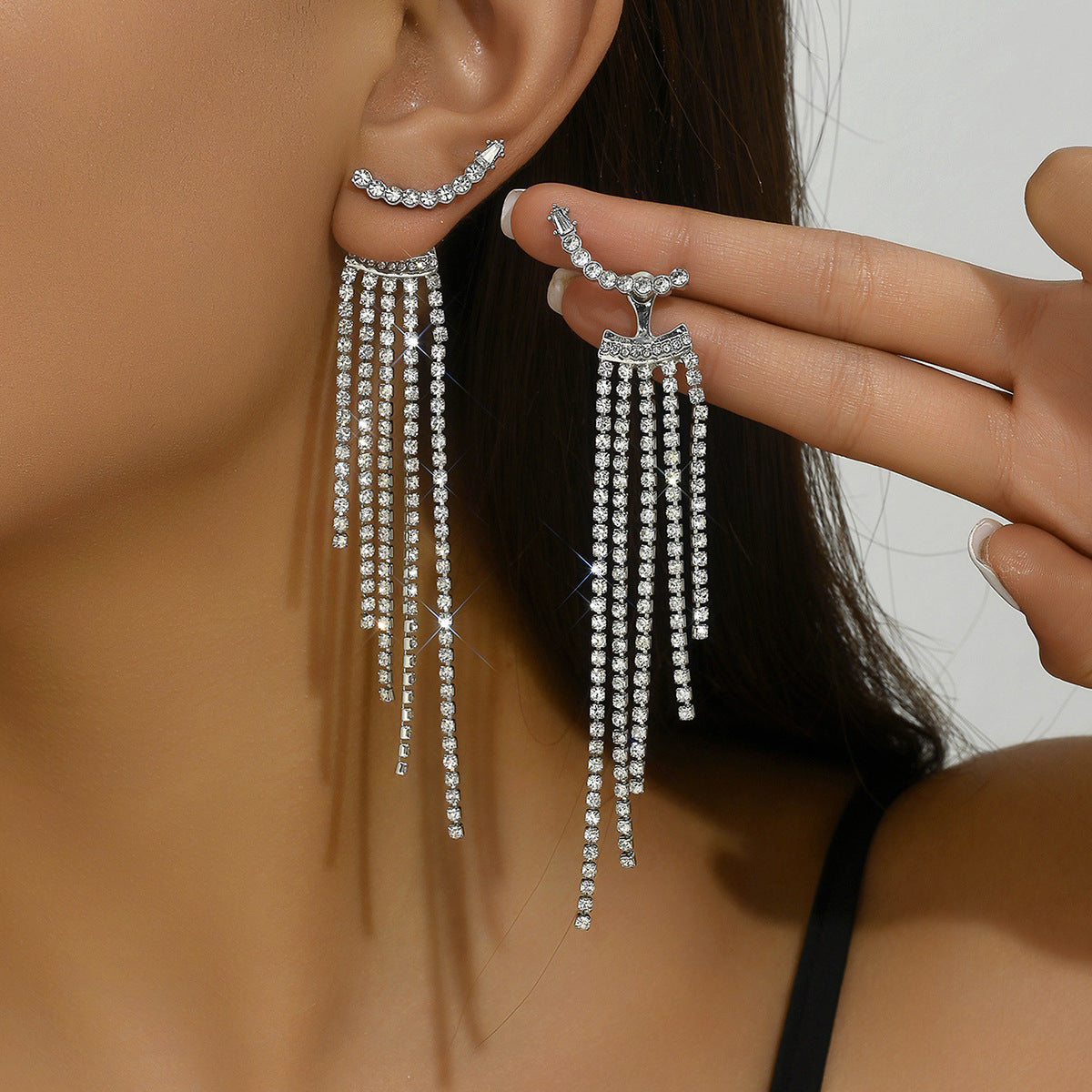A close-up of a model wearing a long, dangling silver-toned statement earring on one ear while holding the matching earring against her finger. The earrings feature a curved top set with small clear stones and multiple cascading fringe strands.
