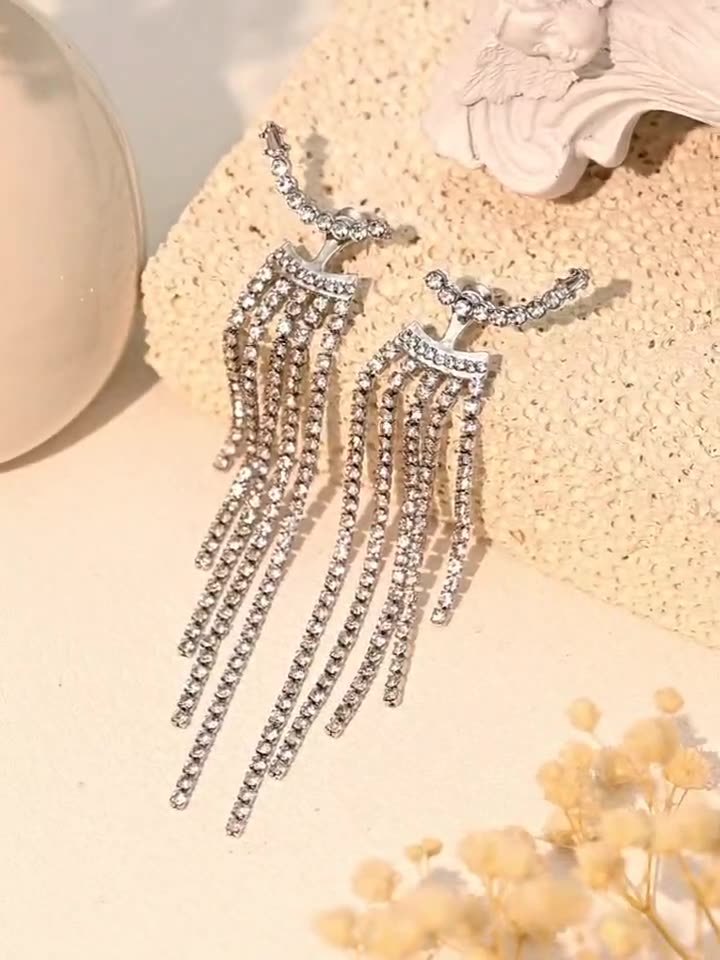 A pair of silver-toned dangle earrings with multiple cascading rows of clear rhinestones, displayed on a textured beige surface with decorative props.