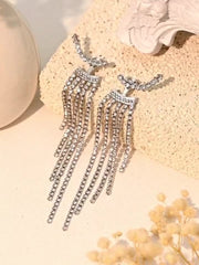 A pair of silver-toned dangle earrings with multiple cascading rows of clear rhinestones, displayed on a textured beige surface with decorative props.