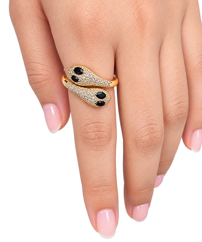 A gold snake ring with black oval eyes and pavé-set clear stones, shown worn on a hand with light pink manicured nails against a plain white background.