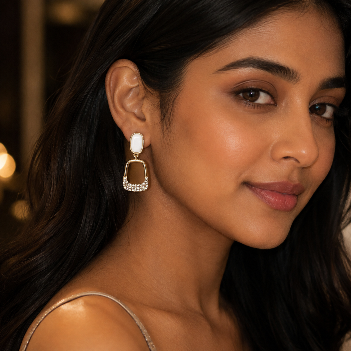 A close-up of a woman wearing a gold-toned dangle earring with a white oval stone and a square frame accented with small clear stones, set against a softly blurred warm-toned background.