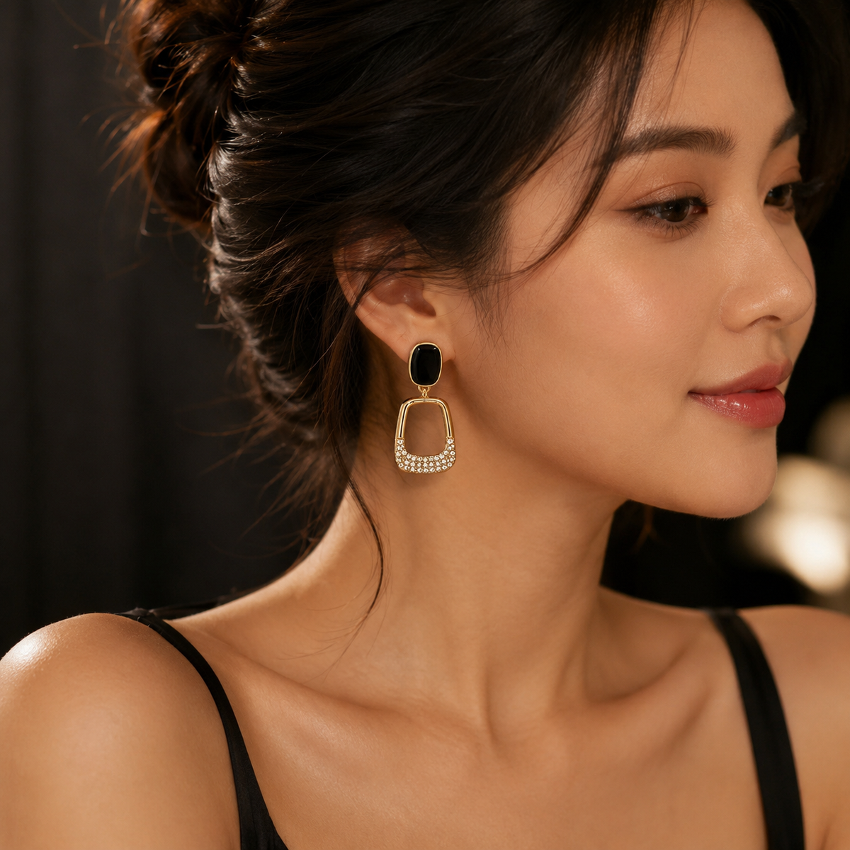 A close-up of a woman wearing a gold-toned drop earring with a black rectangular stone and a dangling square frame set with small clear stones, against a dark blurred background.